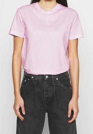 Persoon draagt een lichtroze T-shirt met korte mouwen met een klein wit logo en een zwarte high-waisted spijkerbroek met voorzakken.