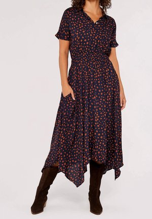 Kobieta ubrana w granatową sukienkę midi w brązowe kropki, z krótkimi rękawami, z ręką w kieszeni, w połączeniu z ciemnobrązowymi kozakami za kolano.