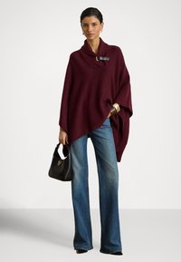 Γυναίκα που φοράει μπορντό πλεκτό πόσο με πόρπη, μπλε τζιν φαρδιάς γραμμής, μαύρη τσάντα χειρός, χρυσά και καφέ βραχιόλια, στέκεται μπροστά σε απλό λευκό φόντο.