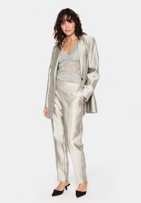 Zilveren satijnen pak met een op maat gemaakte blazer en bijpassende broek. Daaronder een lichtblauwe kanttop. Zwarte puntige hakken maken de look compleet.