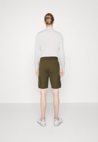 Shorts cargo vert olive avec poches latérales, associés à un haut à manches longues gris clair. Des baskets blanches complètent la tenue.