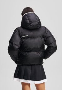 Doudoune noire avec capuche, présentant une texture brillante, un design matelassé et un détail de logo blanc au dos. Associée à une jupe plissée.
