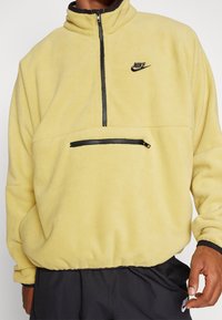 Gul fleecepullover med hög krage, halvdragkedja fram och en ficka med dragkedja på bröstet. Har en svart logotyp och kontrasterande detaljer.