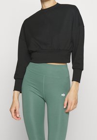 Sudadera negra corta con mangas amplias y una cinturilla ajustada, combinada con leggings verdes de cintura alta que cuentan con un detalle de logo blanco.
