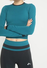 Donna che indossa un crop top a maniche lunghe color turchese e leggings neri con strisce turchesi, braccia conserte, su uno sfondo bianco uniforme.