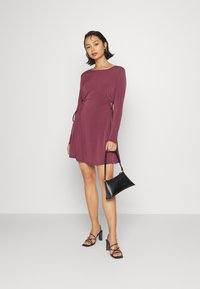 Vestido burgundy de mangas longas com detalhes franzidos na lateral, combinado com uma mini mala preta e saltos bloco com tiras transparentes, de pé contra um fundo branco.