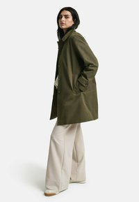 Abrigo verde oliva, de largo hasta la rodilla, con corte recto, bolsillo en el pecho y botones frontales, combinado con pantalones de pierna ancha beige y zapatillas.