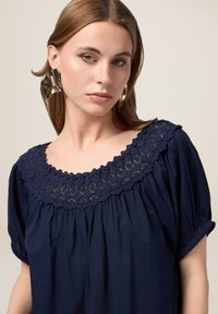Blusa blu navy con maniche a sbuffo corte, caratterizzata da un collo rotondo decorato con dettagli in pizzo. Realizzata in un tessuto morbido, con una vestibilità rilassata.
