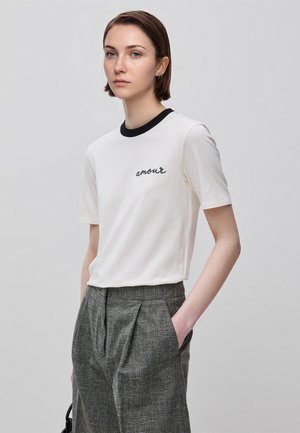 Młoda kobieta z krótkimi brązowymi włosami, ubrana w białą koszulkę z napisem "amour" i szare spodnie w kratę, stoi z jedną ręką w kieszeni.