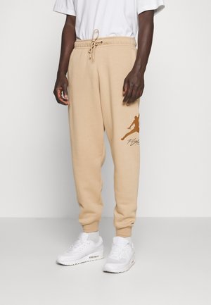 Mørkhudet person iført beige sweatpants med brun Jumpman-logo og hvide sneakers, stående på en ensfarvet hvid gulv.
