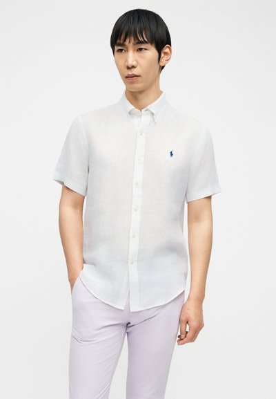 Νεαρός άνδρας που φοράει λευκό πουκάμισο με κοντά μανίκια και κουμπιά, με μπλε κεντημένο λογότυπο, και ανοιχτό μωβ παντελόνι, στέκεται με το ένα χέρι στην τσέπη.