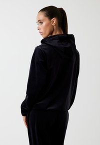 Vrouw met een paardenstaart, gekleed in een zwarte hoodie en bijpassende broek, kijkt naar de zijkant tegen een effen lichtgrijze achtergrond.