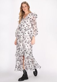 Kleid mit Blumenmuster aus durchsichtiger weißer Stoff mit schwarzen Blumen, Rüscheärmeln, Wickeldesign und stufigem Saum. Kombiniert mit schwarzen Knöchelstiefeln.