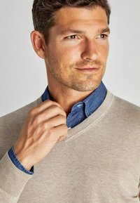 Beige V-Ausschnitt-Pullover, der über einem blauen Jeanshemd getragen wird. Der Pullover hat gerippte Bündchen und eine weiche Textur. Eine Hand berührt den Kragenbereich.