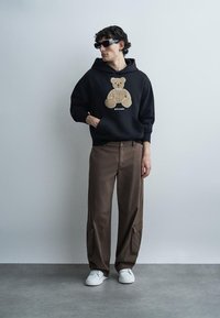 Vīrietis mugurpusē melnā kapucē ar lāča attēlu, brūnas kargo bikses, baltas sporta kurpes un melnas saulesbrilles, stāv pret vienkāršu pelēku sienu.