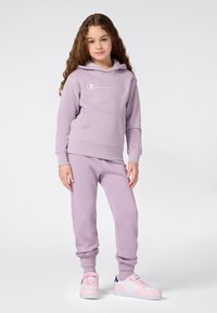 Niña pequeña con cabello rizado que lleva una sudadera de Champion color lavanda y pantalones de chándal a juego, con zapatillas rosas y blancas, de pie contra un fondo blanco.