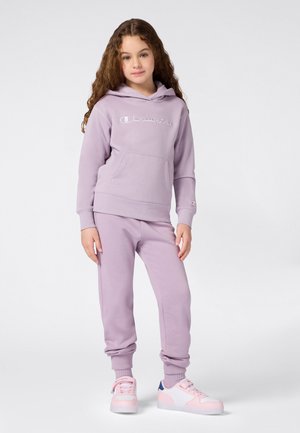 Ragazzina con capelli ricci che indossa un hoodie Champion lilla e pantaloni della tuta coordinati, con sneakers rosa e bianche, in piedi su uno sfondo bianco.