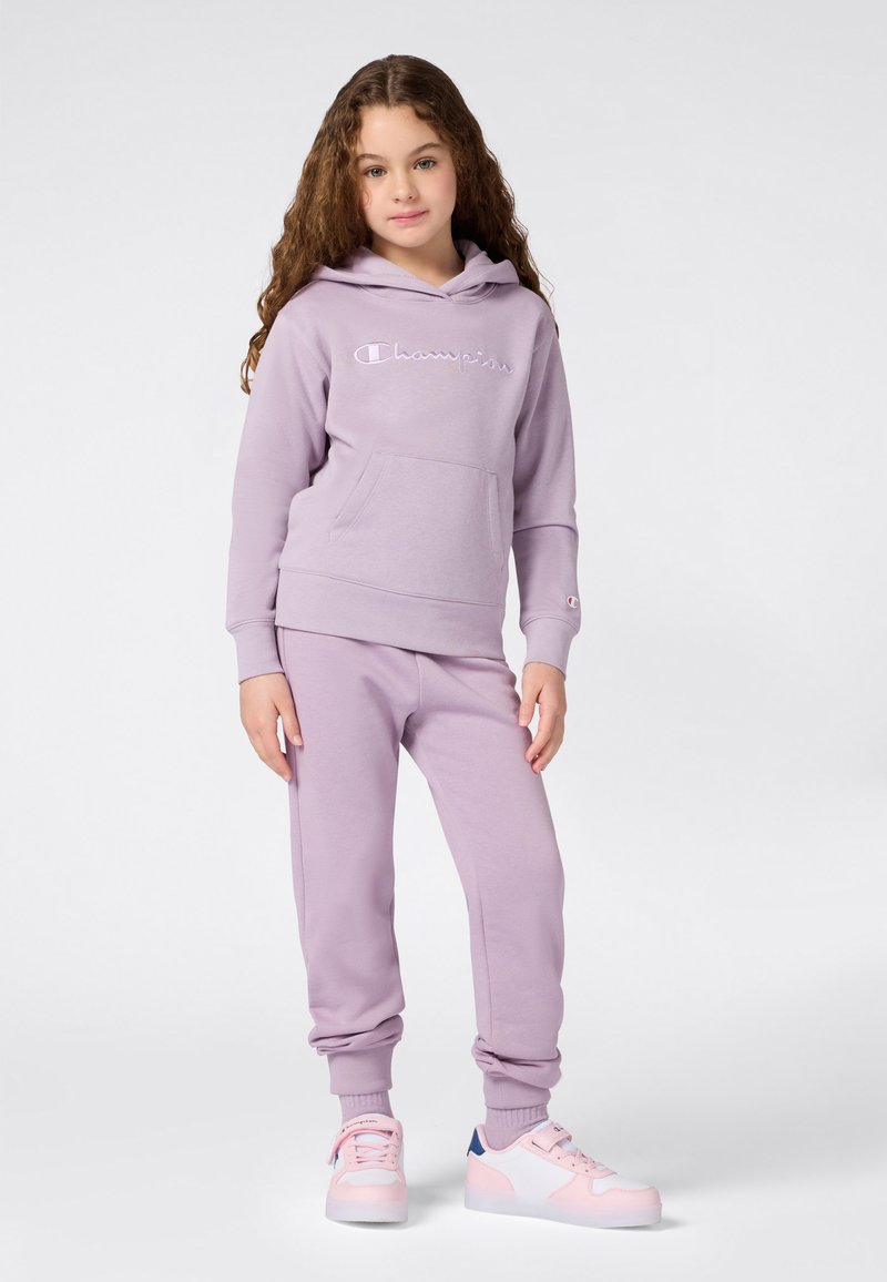 Niña pequeña con cabello rizado que lleva una sudadera de Champion color lavanda y pantalones de chándal a juego, con zapatillas rosas y blancas, de pie contra un fondo blanco.