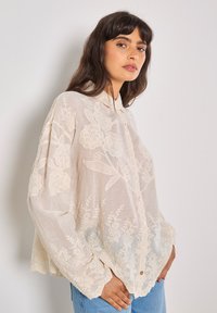 Blusa trasparente di colore crema, con ricami floreali, chiusura a bottoni e colletto. Vestibilità rilassata e dettaglio a fondo scallop.
