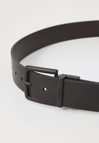 Ceinture en cuir noir avec une sangle plate, dotée d'une boucle rectangulaire en métal noir mat et de perforations régulièrement espacées pour l'ajustement.