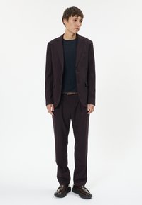 Sötét lila öltöny, szabott blazerrel és passzoló nadrággal, tengerészkék pulóverrel párosítva, barna bőr cipőkkel kiegészítve.