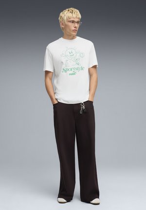 Młody mężczyzna z krótkimi blond włosami, noszący okulary, białą koszulkę z grafiką Puma Sportstyle, czarne szerokie spodnie i białe buty, stojący z rękami w kieszeniach.