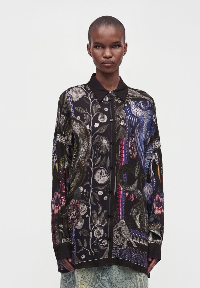 Personne portant une chemise noire à manches longues avec des motifs colorés de serpent, d'oiseau et de fleurs, debout devant un fond clair uni.