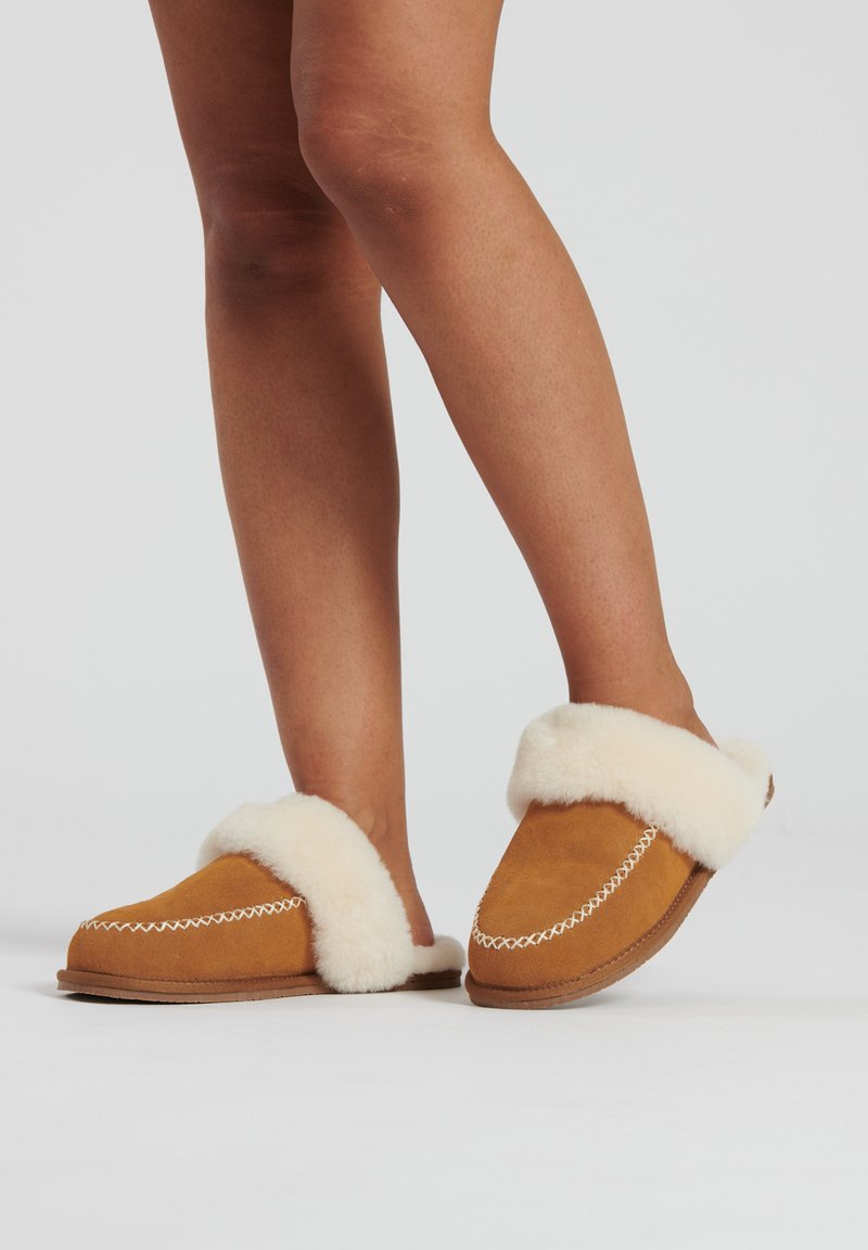 Chaussons en daim marron avec un revers en fausse fourrure crème, dotés de coutures blanches le long des bords et d'une semelle plate.
