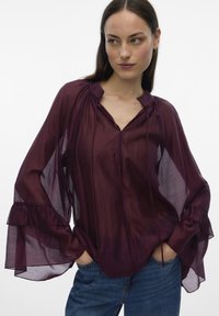 Frau trägt transparente bordeauxrote Bluse mit ausgestellten Ärmeln und Rüschenmanschetten, kombiniert mit blauer Jeans, steht mit den Händen in den Taschen.