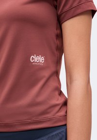 Близък план на човек, носещ ръждиво оцветена тениска Ciele Athletics и тъмносини шорти, показващ логото на гърдите.