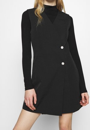 Vrouw draagt een zwarte mouwloze jurk met twee decoratieve knopen over een zwart top met lange mouwen, staand tegen een effen achtergrond.