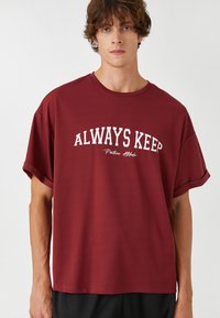 Camiseta corta de manga en burdeos en algodón texturizado, con el texto "SIEMPRE MANTEN" en blanco arqueándose por el pecho. Diseño de corte relajado.