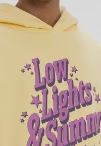 Gelbe Kapuzenjacke mit lila grafischem Text "Low Lights & Sunny." Mit Kordelzugkapuze und Sternenakzenten. Weiches, strukturiertes Material.