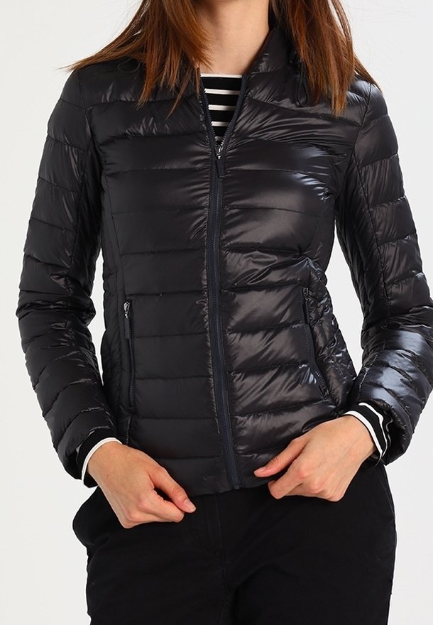Femme portant une veste matelassée noire à fermeture éclair avec poches latérales, associée à un pantalon noir et à une chemise rayée noire et blanche en dessous.