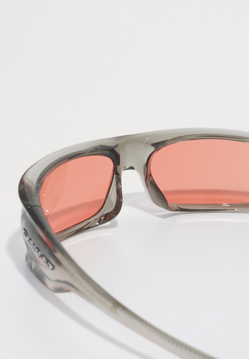 Occhiali sportivi con montatura grigia e lenti rosa. Il design presenta una forma avvolgente e finiture testurizzate sulle aste.