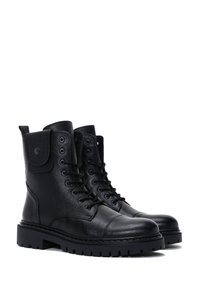 Par de botas de combate de cuero negro con cordones, suelas de goma gruesas y un pequeño bolsillo abotonado en el lado exterior de cada bota.