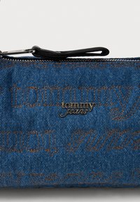 Denimová taška s modrým textilným povrchom, s vyrazeným logom 'tommy' a hnedými švami, uzatvorená zipsom a čiernym ťahákom.