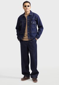 Mørk denimjakke med to brystlommer og gyldent syning, parret med en kabelstrikket tan sweater, løse mørke jeans og brune sko.