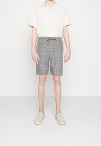 Pantaloni corti a quadri grigi con vita elastica e lacci. Indossati con una camicia beige a maniche corte con colletto e scarpe di colore chiaro.