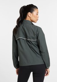 Casaco leve de um verde escuro com colarinho levantado, detalhe refletor nas costas e punhos elásticos para uma silhueta ajustada.