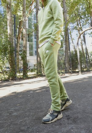 Personne portant un survêtement Nike vert clair et des baskets noir et blanc, debout sur un chemin de terre à côté d'une clôture et des arbres.