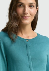 Turkusowy dzianinowy kardigan z zaokrąglonym dekoltem, ozdobiony złotym guzikiem przy kołnierzu i gładką fakturą.