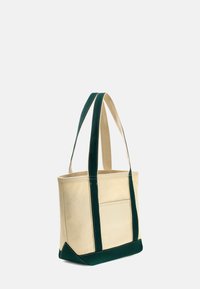 Borsa tote in tela di colore naturale con manici e fondo verde scuro. Presenta cuciture verticali e una tasca esterna su un lato.