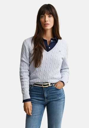 Kvinde iført en lysegrå kabelstrikket V-hals sweater over en mørkeblå skjorte med knapper, blå jeans og et sort-hvidt bælte, stående med den ene hånd i lommen.