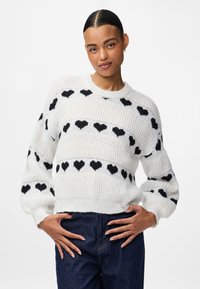 Ung kvinna med mörkt hår i en knut, iklädd en vit stickad tröja med svarta hjärtmotiv, står med händerna på höfterna mot en vit bakgrund.