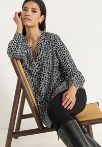 Svartvit mönstrad blus med V-hals och långa ärmar, i kombination med svarta leggings och knähöga svarta stövlar, sittande på en träl stool.