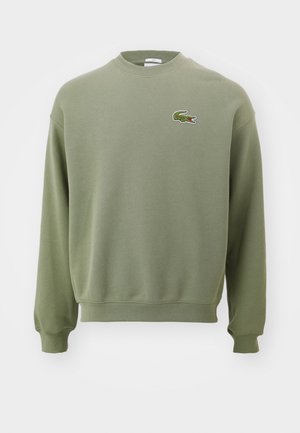 Olive zaļš džemperis ar apaļu kaklu, ribotām piedurknēm un apakšmalu. Uz kreisā krūtis izšūta krokodila logotips. Mīksta, teksturēta auduma.