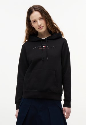 Νεαρή γυναίκα με μακριά καστανά μαλλιά, που φοράει μαύρο φούτερ Tommy Jeans και σκούρα φούστα, στέκεται μπροστά σε λευκό, απλό φόντο.