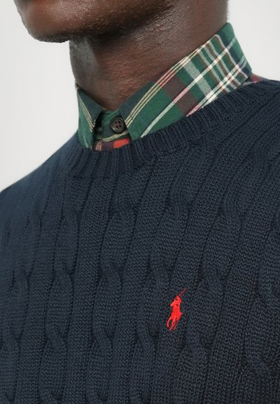 Hombre vistiendo una camisa de cuadro verde con botones debajo de un suéter de punto azul marino con un pequeño logotipo bordado en rojo en el pecho.