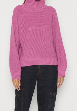 Fuksjowy sweter z wysokim kołnierzem, z dzianiny o fakturowanym wzorze i ściągaczach, zestawiony z ciemnymi spodniami typu cargo.